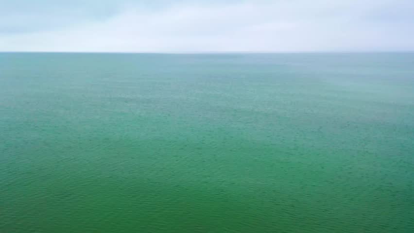 Low-altitude drone footage gliding above calm ocean waters under a cloudy sky, capturing gentle ripples, green-blue tones, and a wide horizon that fades into misty atmospheric light.
