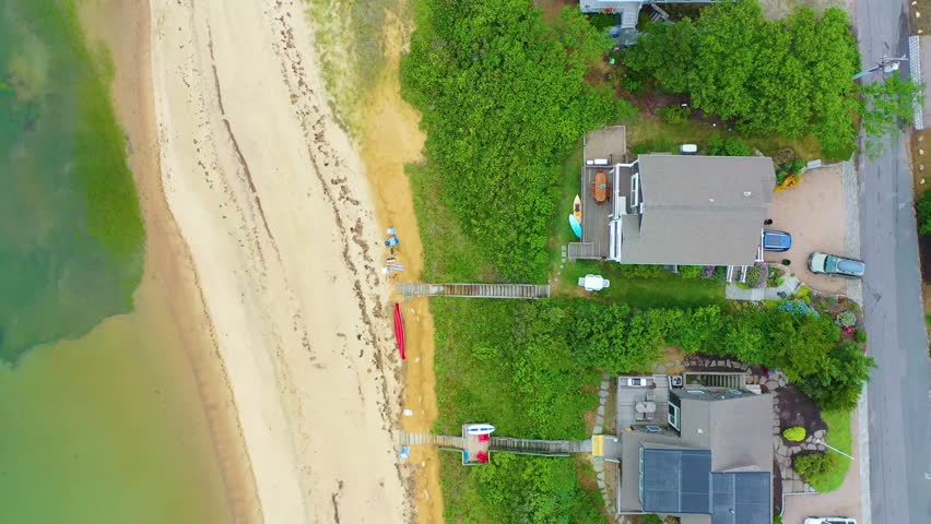 Scenic top-down drone view of Cape Cod shoreline homes, sandy beach, ocean waves, and surrounding greenery, showing neighborhood roads and walkways to the waterfront.
