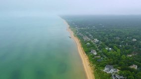 Aerial footage of Cape Cod beach houses on a misty morning, with calm ocean waves, sandy shoreline, cloudy skies, and neighborhoods surrounded by greenery blending into the foggy horizon. - Powered by Shutterstock - Get 15% off with code: PIKWIZARD15