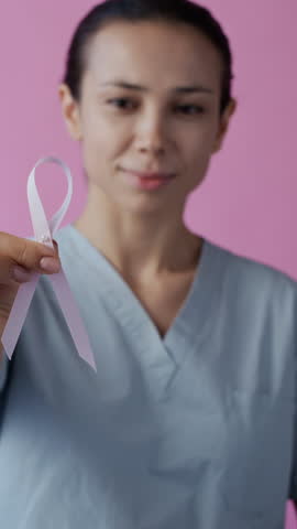 Vertical portrait of young Caucasian female doctor placing pink breast cancer awareness ribbon on medical uniform and looking at camera against pink background