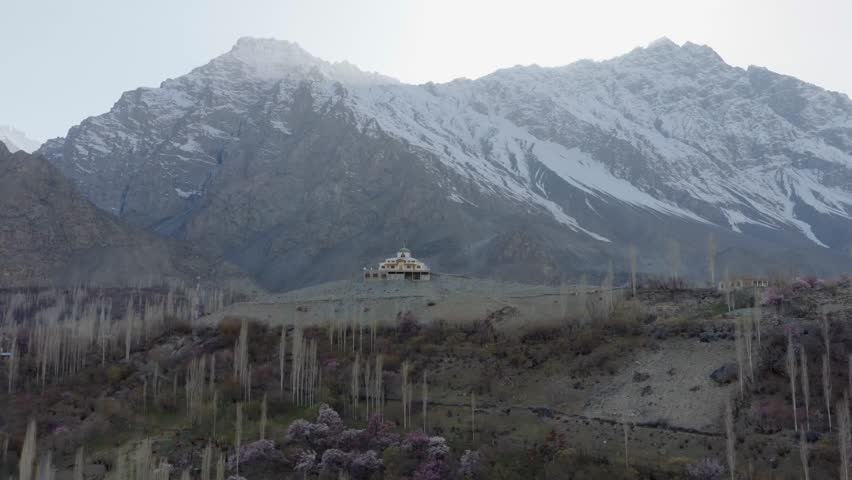 Aerial view of a serene valley in Pakistan, featuring a white building with a dome-like structure against a backdrop of majestic snow-capped mountains