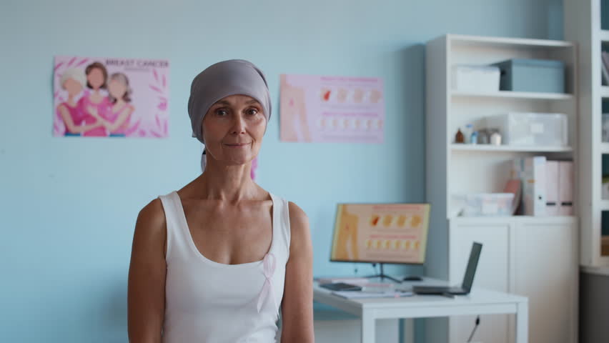 Medium portrait of middle-aged Caucasian woman with breast cancer wearing headscarf and pink ribbon looking at camera in clinic, copy space