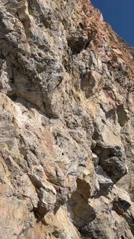 A rocky cliff face with various textures and colors. The surface shows layers of stone and minerals under a clear blue sky.
