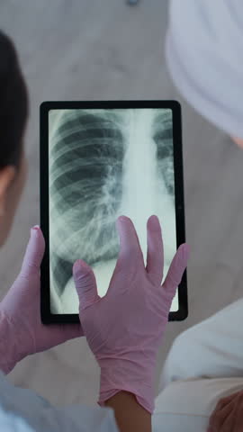 Vertical shot of doctor in medical gloves examining x-ray image on digital tablet sitting next to female patient with cancer in clinic