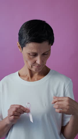 Vertical portrait of mature Caucasian woman with short dark haircut placing pink ribbon on white t-shirt symbolizing breast cancer awareness on pink background