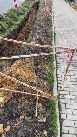 A trench has been excavated, bordered by wooden barricades and vibrant warning tape, positioned along a publicly accessible sidewalk