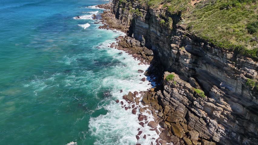 Aerial 4K drone footage of dramatic rocky cliffs meeting the turquoise Pacific Ocean on the New South Wales coast of Australia.