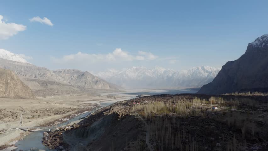 Aerial view of a serene valley in Pakistan, featuring a river flowing through a rocky landscape with snow-capped mountains in the background