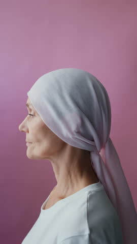 Vertical portrait of mature Caucasian woman with cancer in remission taking off headscarf looking at camera isolated on pink