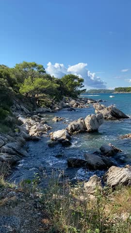 Rocky coastline with trees and bushes under blue sky with clouds, ocean waves hitting shore video