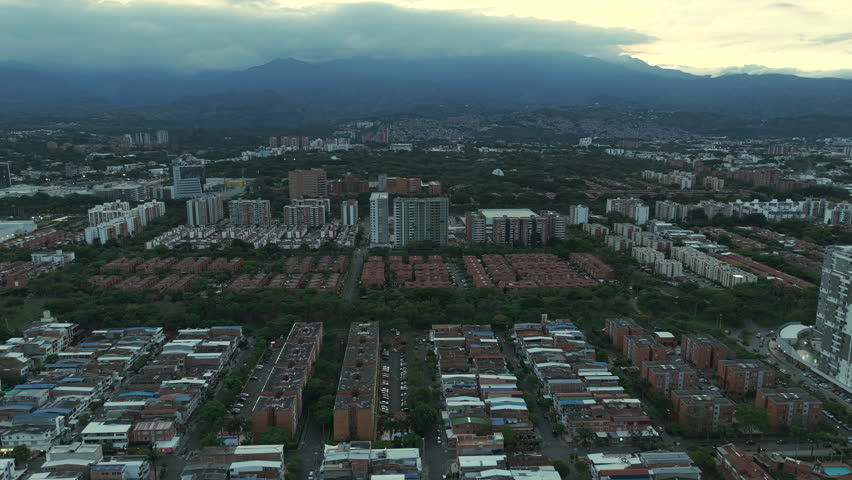 Drone captures the urban sprawl of Valle del Lili, Cali, under a dramatic cloudy sky at dusk. The expansive view showcases residential buildings and lush greenery.