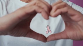 Zoom out shot of anonymous woman making heart shape with hands around breast cancer awareness pink ribbon wearing white t-shirt isolated on pink background - Powered by Shutterstock - Get 15% off with code: PIKWIZARD15