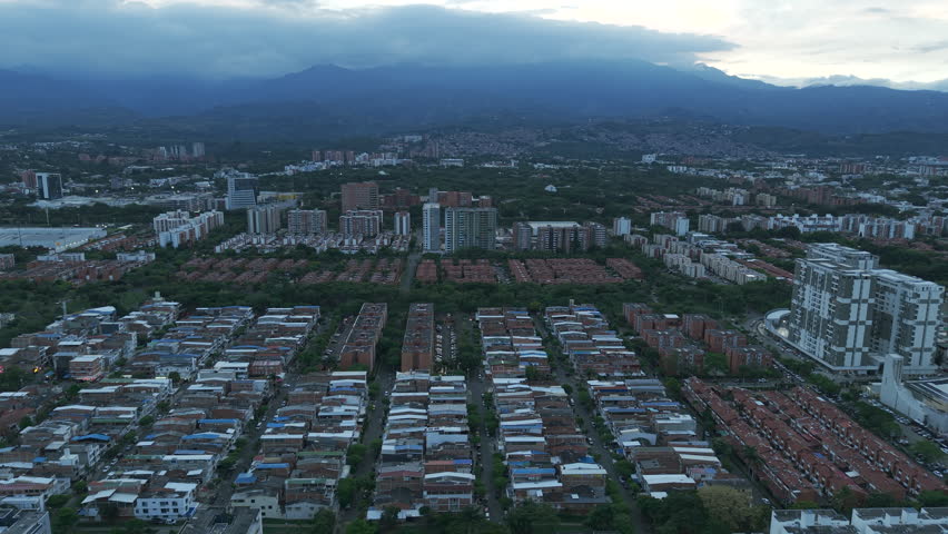 Drone footage captures the sprawling urban landscape of Valle del Lili, Cali at dusk, with rows of buildings, lush greenery, and majestic mountains under a dramatic sky.