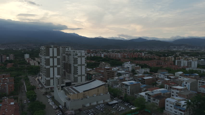 Drone video over Cali's Valle del Lili, showcasing urban sprawl under a dramatic sunset. The sky's warm hues contrast with towering buildings, creating a captivating visual.