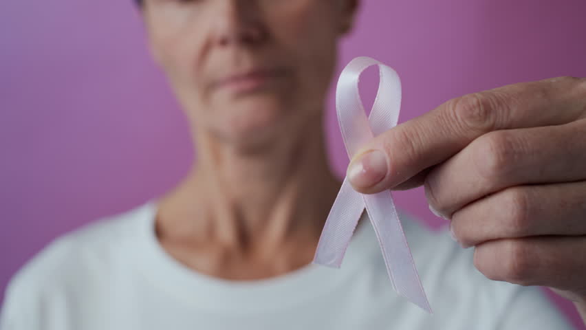Rack focus of mature Caucasian woman, breast cancer survivor, holding pink ribbon looking at camera with smile on pink background, female solidarity concept