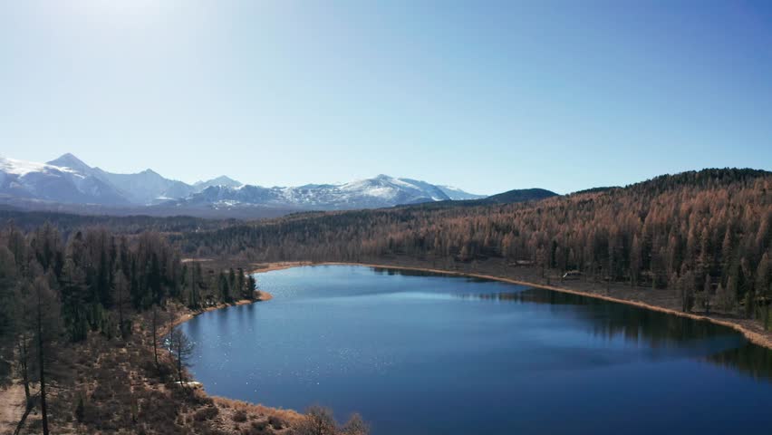 Autumn view of blue mountain lake with sunlight reflecting on surface of lake, snow-capped mountain peaks on background. Forward Flight approach drone shot of Lake Kidelyu, Ulagansky Track, Altai