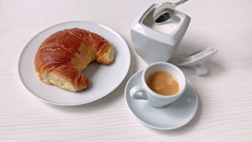 Male hand pouring and stirring sugar into coffee cup. Top view of espresso and croissant breakfast on light wooden table.