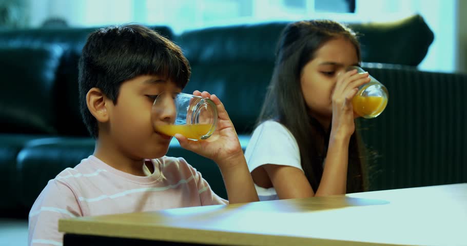 Indian asian small kids drinking juice in glass at home indoors, young brother sister siblings enjoying fruit juice or cold drink together, healthy lifestyle diet with family bonding in modern home