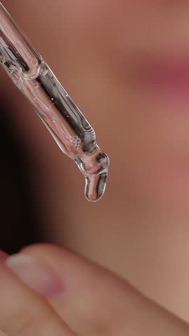 VERTICAL VIDEO: Young woman holding a dropper with serum for face. Facial oil fall from a glass pipette, extreme close-up. Cosmetic backdrop with liquid skincare product. Beauty concept. Slow motion.