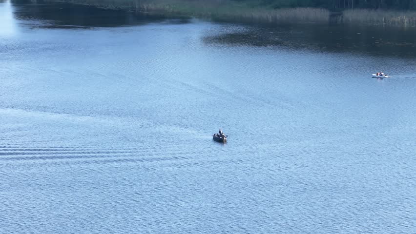 Aerial view of two small boats with people enjoying a ride on a calm lake, leaving rippling trails across the reflective blue water surface.

