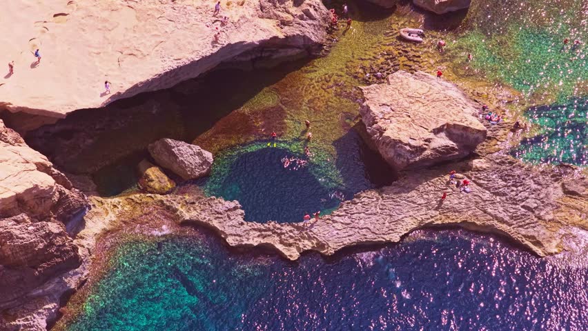 4K drone soars over Gozo’s Blue Hole at midday: deep circular pool, turquoise shallows, swimmers and dramatic limestone edges—perfect for travel, nature, conservation, tourism and documentary use.