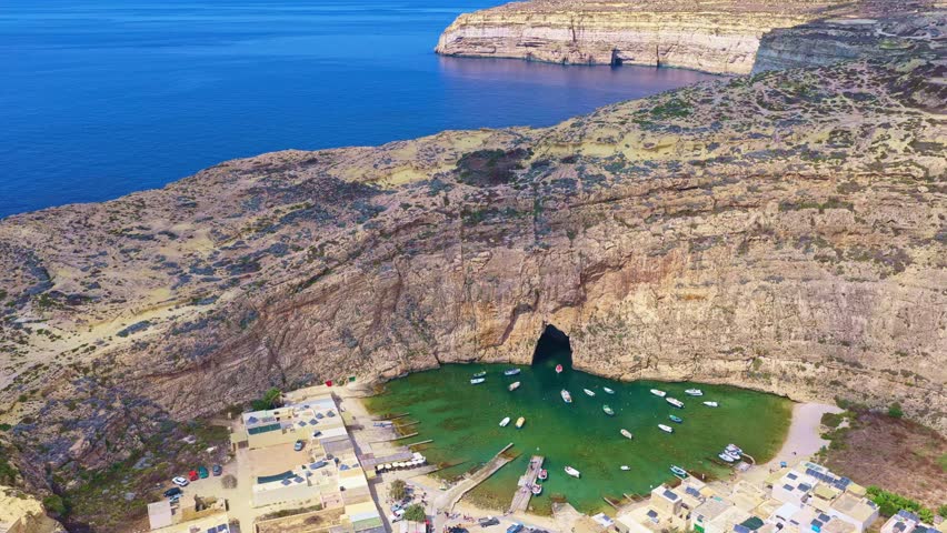 4K drone circles Gozo’s Inland Sea at midday: emerald lagoon, towering limestone cliffs, cave passage to the open sea and moored boats—ideal for travel, geology, tourism, conservation, and documentary
