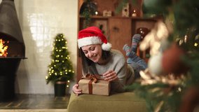 Merry Christmas. Young woman in Santa hat unpacking open up gift box near Christmas tree. Girl in living room with Christmas tree, fireplace opening gift box with surprise face. Christmas eve at home - Powered by Shutterstock - Get 15% off with code: PIKWIZARD15