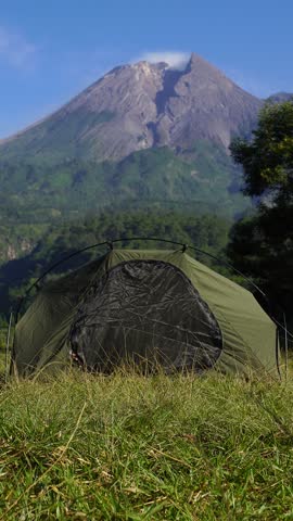 A male adventurer goes solo camping, setting up a tent in the grass with a beautiful volcano in the background. Outdoor activity: setting up a tent around an active volcano on a hot morning.