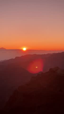 Spectacular sunset over the volcanic mountains of Gran Canaria, with glowing orange horizon, misty valleys and dramatic golden hour light creating a breathtaking natural landscape.