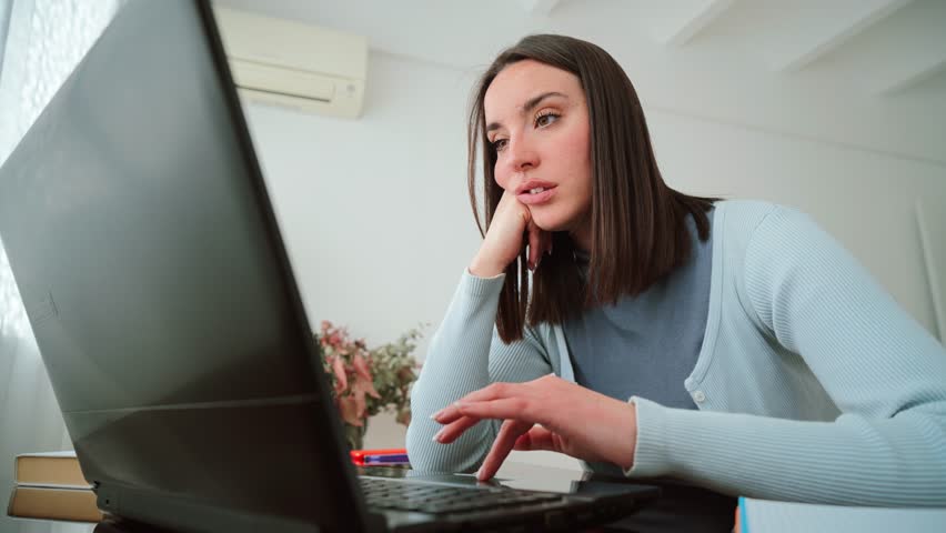 A young woman looking frustrated while working on her laptop at home, showing signs of sadness and stress on her face as she attempts to concentrate on her studies and tackle her academic tasks