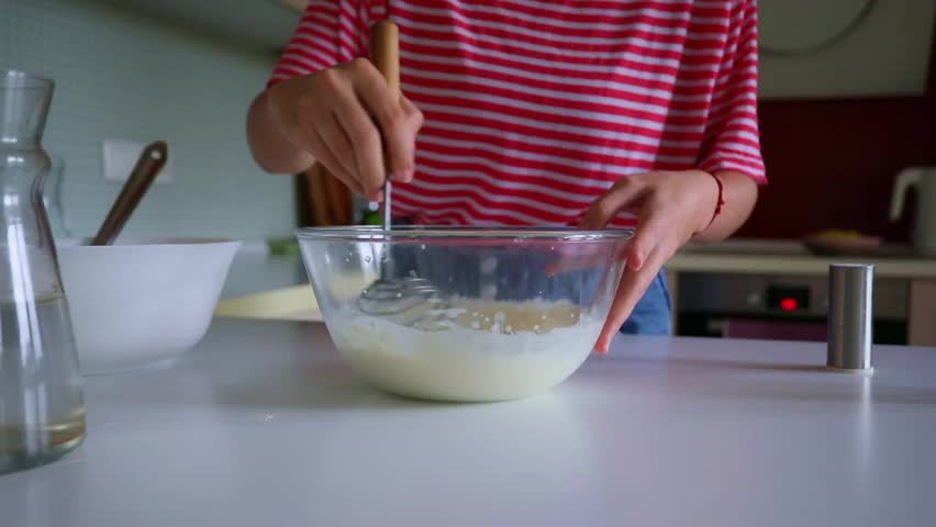 Close-up of a person in a red striped shirt whisking batter in a glass bowl on a kitchen counter, preparing homemade food.