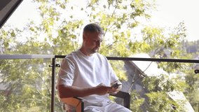 Casual man in white clothes sitting on balcony with coffee cup and smartphone, enjoying peaceful outdoor view of trees and houses. Concept of relaxation, modern lifestyle, and digital connection. - Powered by Shutterstock - Get 15% off with code: PIKWIZARD15