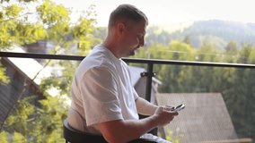 Casual man in white clothes sitting on balcony with coffee cup and smartphone, enjoying peaceful outdoor view of trees and houses. Concept of relaxation, modern lifestyle, and digital connection. - Powered by Shutterstock - Get 15% off with code: PIKWIZARD15