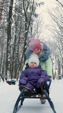 in the snow-covered winter forest, two girls, one year and seven years old, have fun sledding. the elder sister rolls the younger sister on the sled