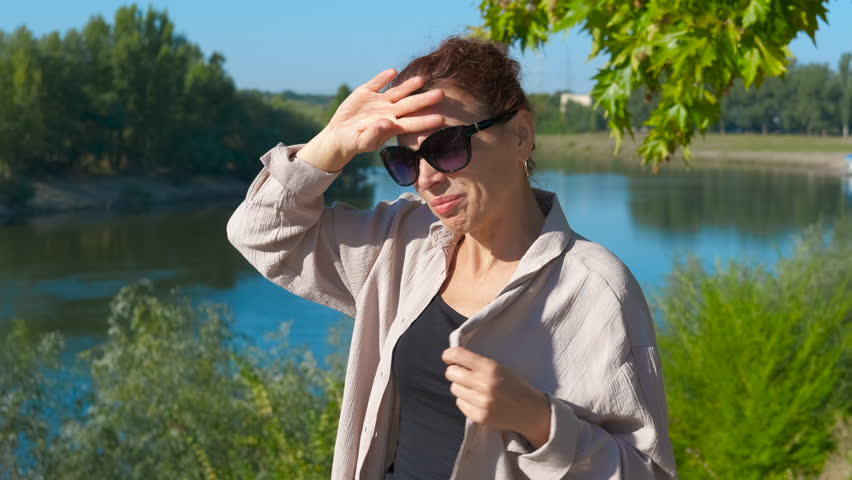 Woman suffering from summer heatstroke near river. Mature woman experiencing heatstroke symptoms while standing near river, pressing hand against forehead and readjusting clothing during intense heat