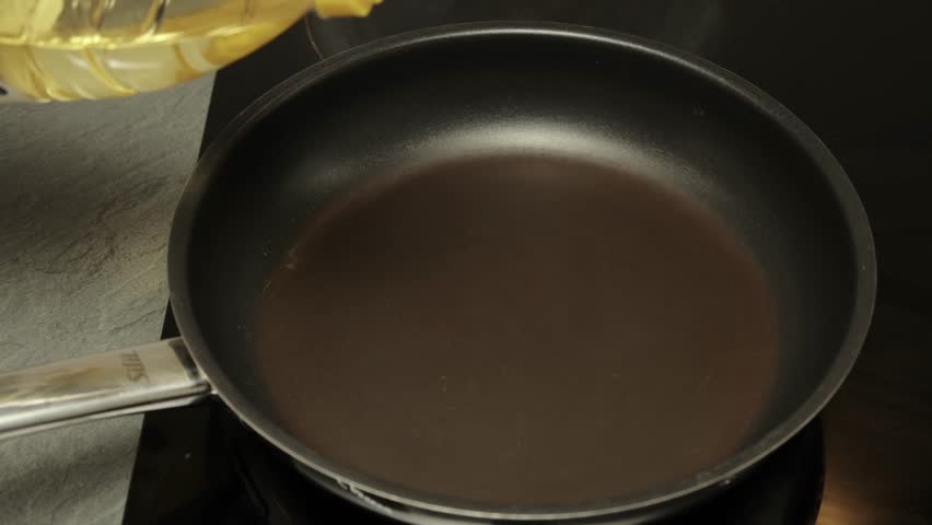 Female hand pours sunflower oil into a frying pan. Bright top view captures the cooking preparation step. Minimalist kitchen scene with natural light on white background.