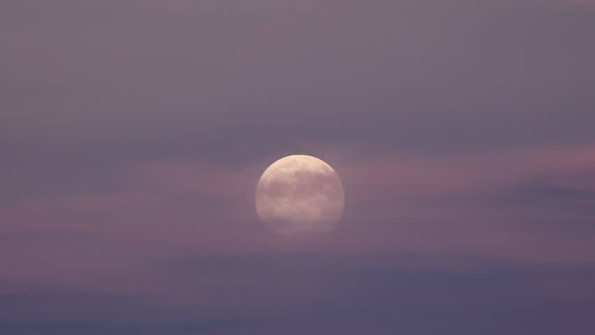 A 99 percent full moon slowly rises from behind thin dark mauve clouds that drift from right to left in the evening sky just after sunset on a September evening.
