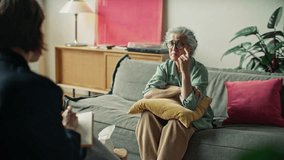 Sad senior woman sitting on sofa, holding yellow pillow and covering face in distress while crying during therapy session with female psychologist in cozy modern office - Powered by Shutterstock - Get 15% off with code: PIKWIZARD15