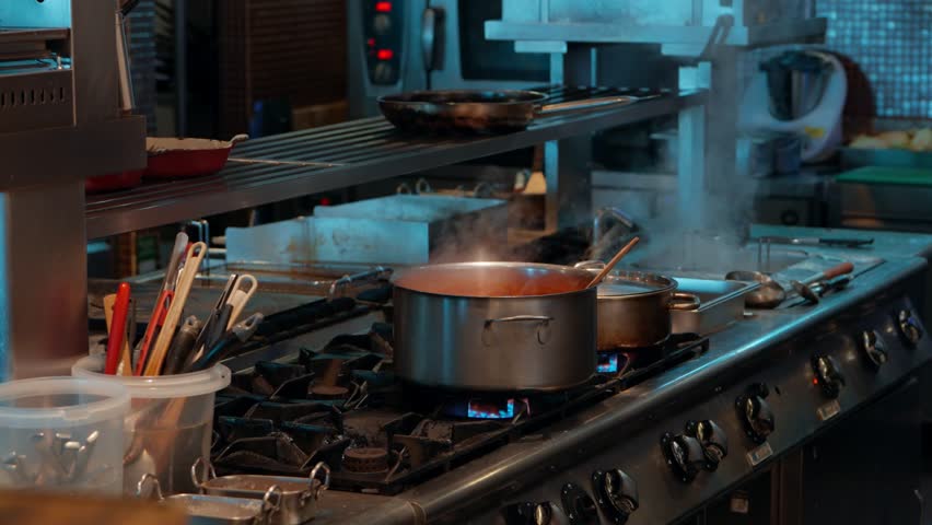 A wide shot of a busy professional restaurant kitchen showing a large pot of sauce simmering on a stainless steel gas stove while a chef works in the background, a behind-the-scenes culinary view