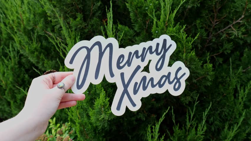 Young Woman Holds Up message of Merry Christmas Wording in front of Christmas Tree in a garden in Outside 4K Video Stock Footage