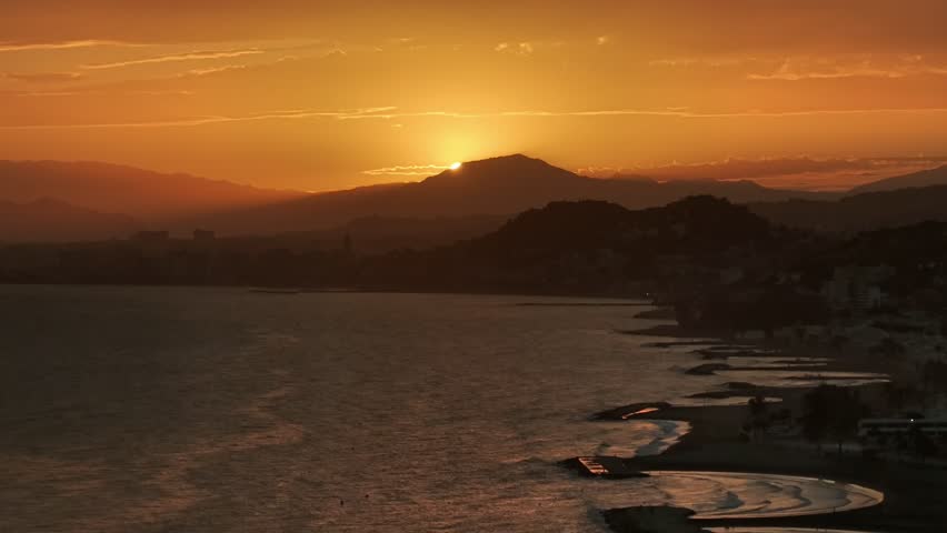 Golden sunset over Málaga’s coastline, with the sun dipping behind mountains, casting warm light across the sea and silhouetted cityscape.