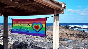Slow motion of a colorful rainbow flag with the word love and a heart waving in the wind under a wooden shelter, with the blue ocean and a volcanic rocky shore in the background - Powered by Shutterstock - Get 15% off with code: PIKWIZARD15