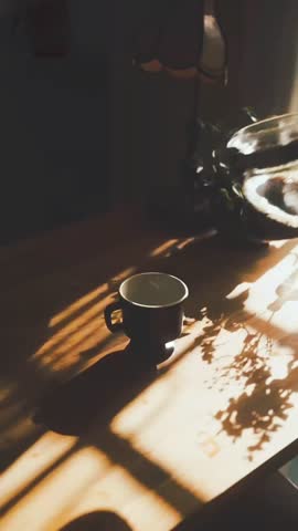 A warm cup of coffee being poured in soft morning sunlight, with shadows of leaves and a cozy atmosphere on a wooden table.