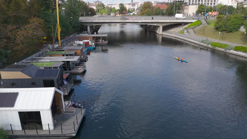 Aerial view of the Brda River flowing under a bridge, lined with houseboats, with kayakers paddling on the dark water, Bydgoszcz, Kuyavian-Pomeranian Voivodeship, Poland.
