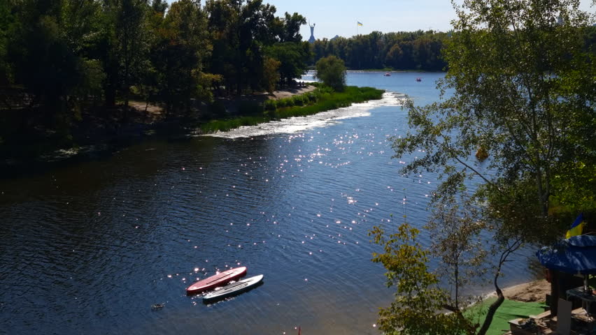 Beautiful summer day on the Dnipro river in Kyiv, Ukraine, with sparkling sunlight reflecting on the water surface and two kayaks floating near the shore, surrounded by green trees