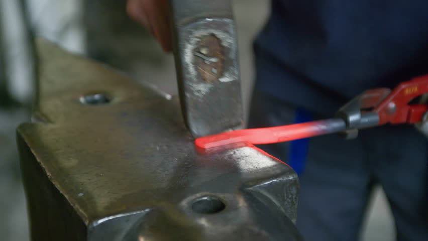 Close-up side view of blacksmith hammering hot glowing metal on anvil. Sparks fly as traditional forging technique and manual precision come to life in the workshop.