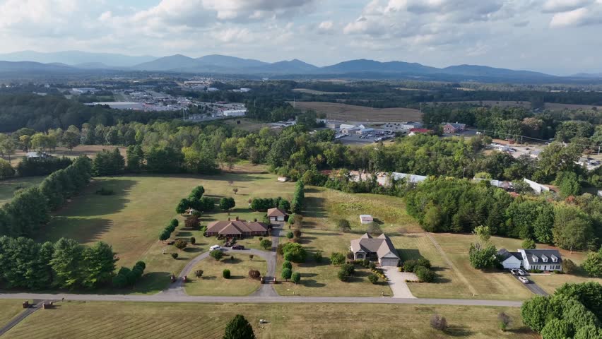 Drone view of American countryside with scattered houses, open fields and distant blue mountains under cloudy sky. Rural lifestyle and wide landscapes in Virginia, USA. Aerial wide shot.