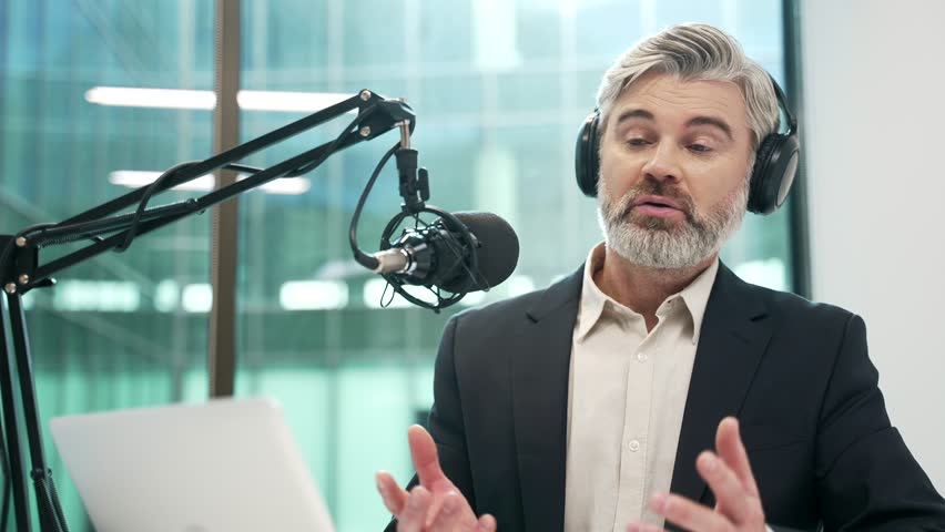 Businessman in wireless headphones recording podcast using microphone and laptop sitting in business office. Tutor giving video call training. Blogger is streaming. Coach has online course. Close up