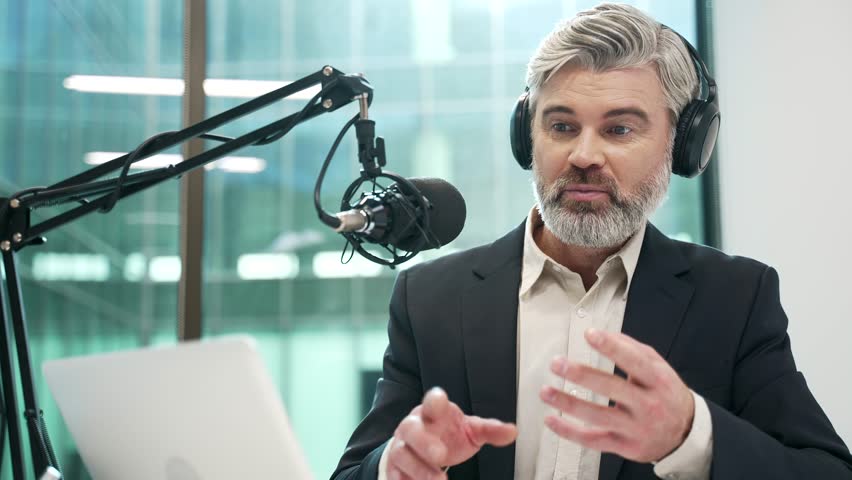 Businessman in wireless headphones recording podcast using microphone and laptop sitting in business office. Tutor giving video call training. Blogger is streaming. Coach has online course. Close up