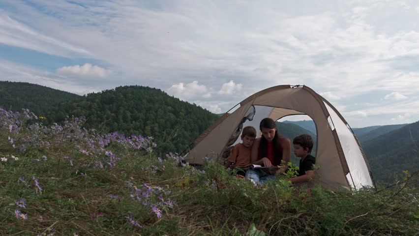 Family gathered inside a tent on a hillside, enjoying a storytelling session, surrounded by wildflowers and mountains, creating a serene outdoor experience for bonding and learning